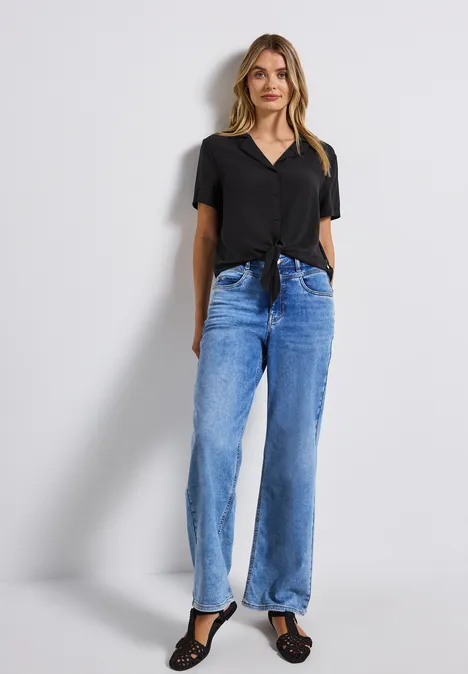 Frau in schwarzer Bluse und weiten Jeans vor weißer Wand.
